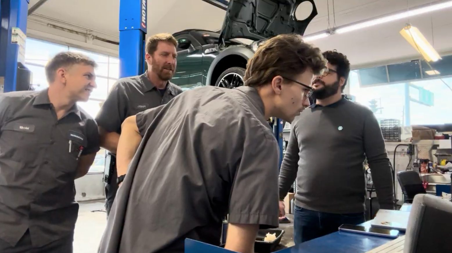 Research session inside a repair shop: technicians in uniform gathered around a workstation with a car raised on a lift behind them